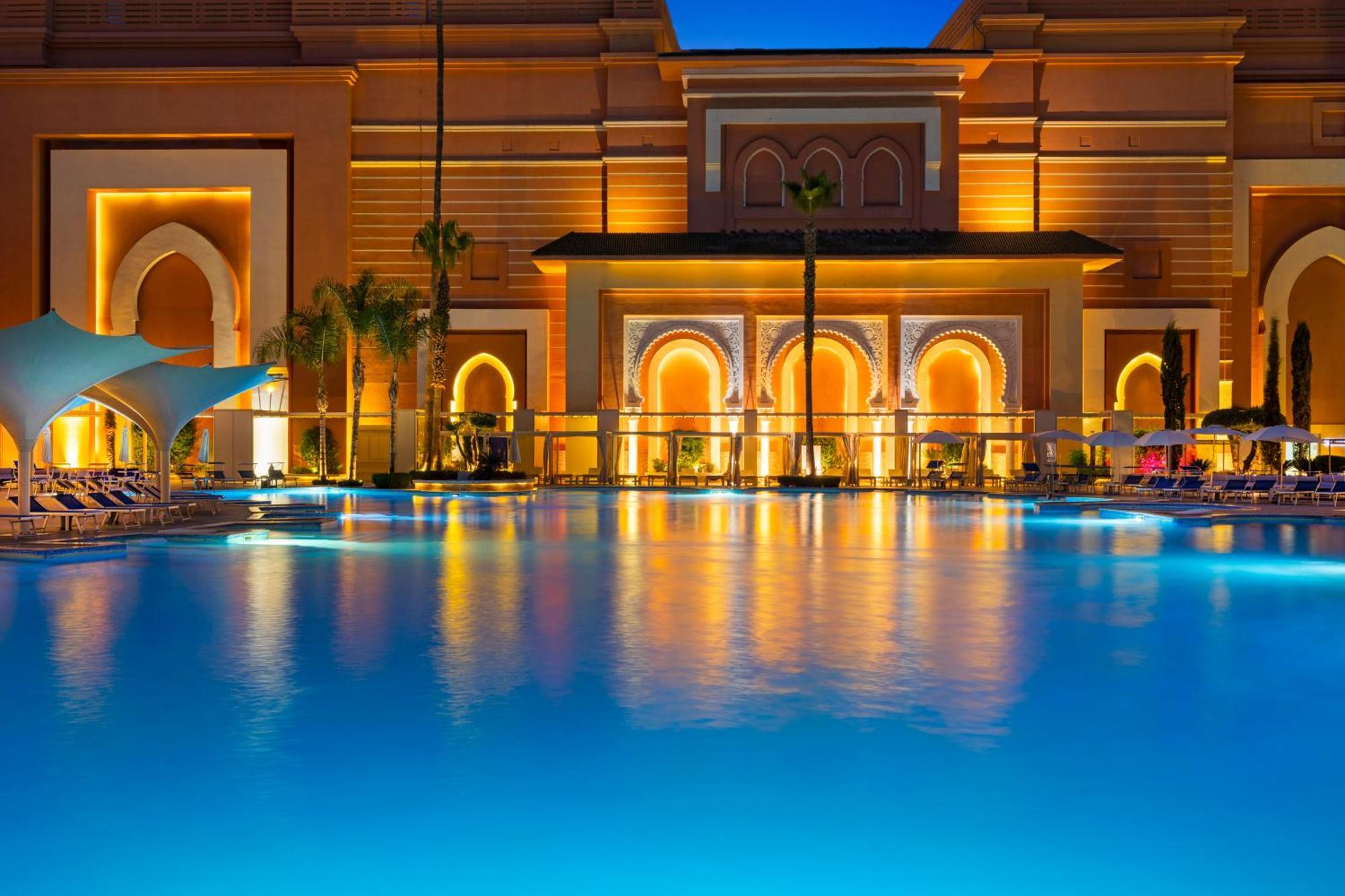 ซาวอย เลอ กร็อง โฮเต็ล มาราเกช Hotel มาร์ราเกช ภายนอก รูปภาพ The photo shows a beautifully illuminated resort at dusk. In the foreground, a swimming pool reflects the blue of the water and the warm lighting of the surrounding architecture. The buildings in the background feature intricate designs with arched d