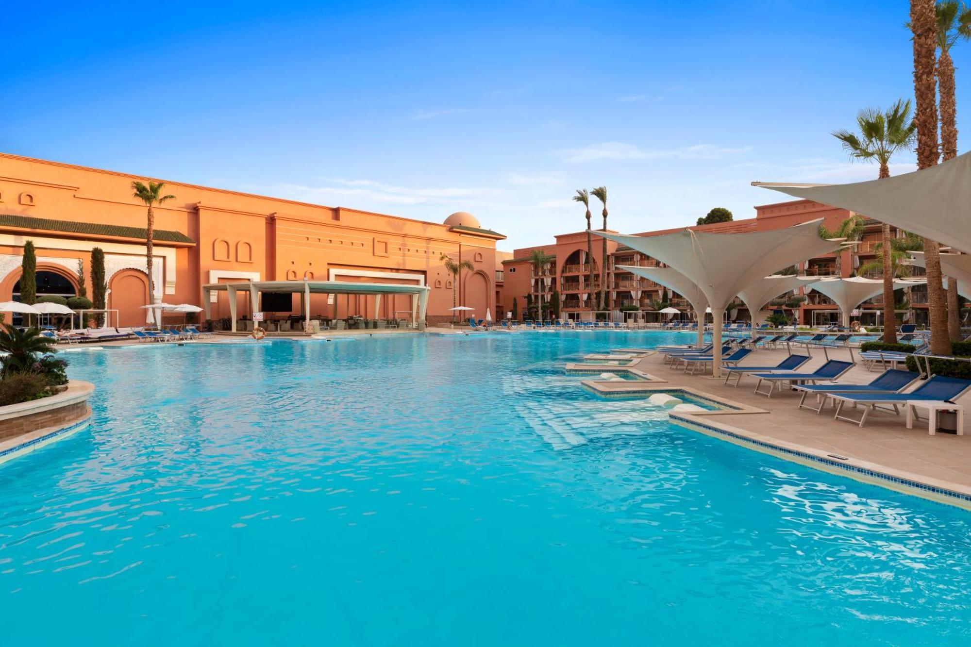 ซาวอย เลอ กร็อง โฮเต็ล มาราเกช Hotel มาร์ราเกช ภายนอก รูปภาพ The photo shows a large outdoor swimming pool with clear, turquoise water. Surrounding the pool are several lounge chairs, some of which are sheltered by large umbrellas. In the background, there are buildings with a distinctly warm-colored architect