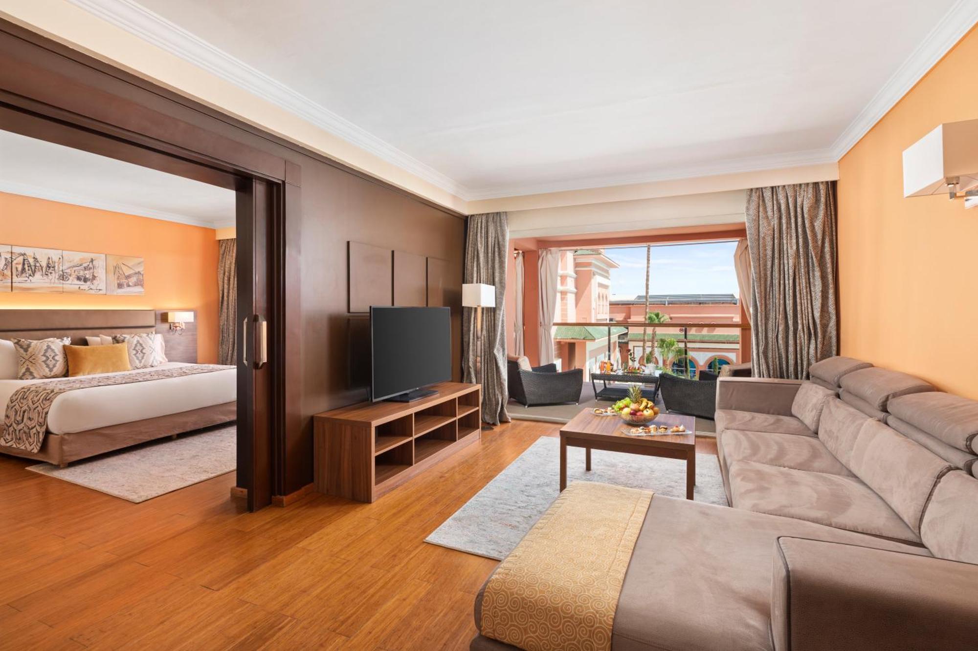 ซาวอย เลอ กร็อง โฮเต็ล มาราเกช Hotel มาร์ราเกช ภายนอก รูปภาพ The photo shows a hotel suite that features a welcoming and modern design. In the foreground, there is a cozy living area with a comfortable beige sofa and a coffee table. A television is mounted on a wooden stand, adding to the entertainment options