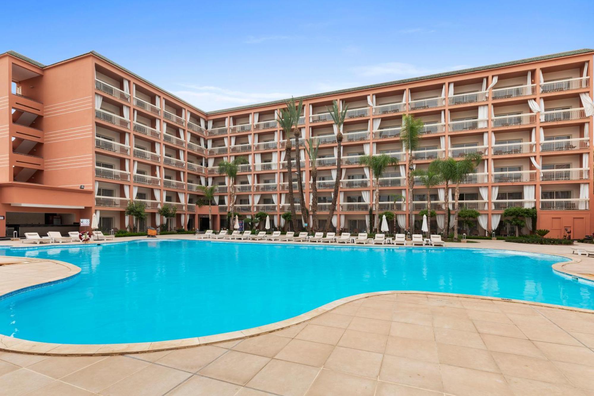 ซาวอย เลอ กร็อง โฮเต็ล มาราเกช Hotel มาร์ราเกช ภายนอก รูปภาพ The photo shows a large swimming pool surrounded by a paved area with lounge chairs. In the background, there is a multi-story building with a pink exterior featuring multiple balconies. The setting appears to be sunny, and there are palm trees nearb