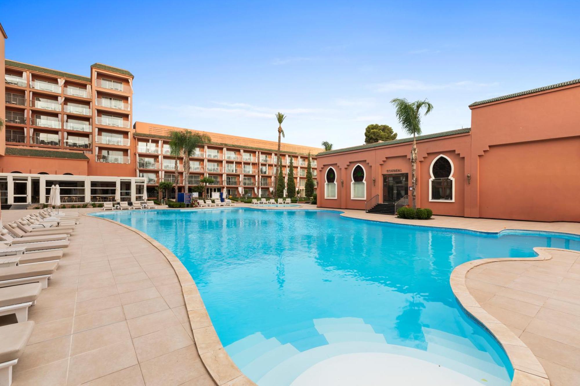 ซาวอย เลอ กร็อง โฮเต็ล มาราเกช Hotel มาร์ราเกช ภายนอก รูปภาพ The photo shows a large swimming pool in an outdoor setting, surrounded by palm trees. On one side of the pool, there is a modern building with multiple floors and balconies, while on the opposite side, there is a structure with a distinctive archite